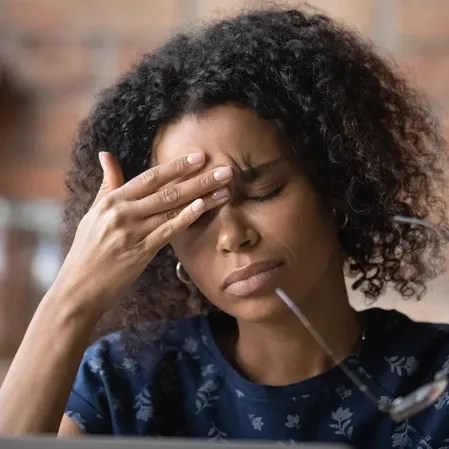 Person touching the forehead while sitting indoors, with eyeglasses visible in the foreground.